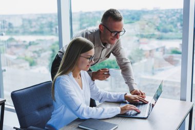 Business woman with a laptop for a male colleague who looks into an open notebook. Business cooperation and teamwork. European millennials at the table in the office. Modern successful people