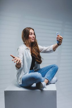 Uzun esmer saçlı, beyaz gömlek ve kot pantolon giyen, telefonda video konuşması yapan genç bir kadın. Kot pantolonlu ve cep telefonu olan beyaz gömlekli bir kadın. Beyaz arkaplan