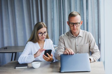 Cheerful businesspeople using a laptop in an office. Happy young entrepreneurs smiling while working together in a modern workspace. Two young businesspeople sitting together at a table.