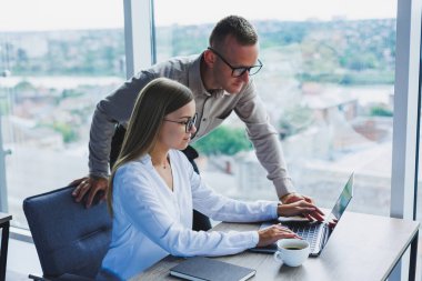 Business woman with a laptop for a male colleague who looks into an open notebook. Business cooperation and teamwork. European millennials at the table in the office. Modern successful people