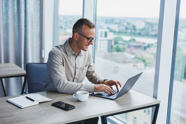 A businessman works on a laptop, a manager sits at a table in the office, works on a laptop. Freelancer workplace, employee at remote work. Remote workplace in coworking, cafe