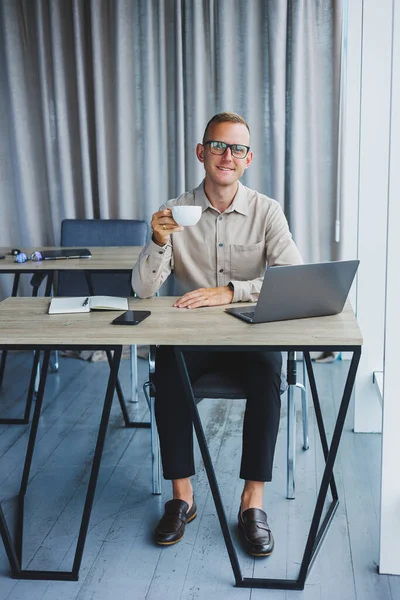 A freelancer in glasses works on a laptop and drinks coffee, a manager sits at a table in the office, works on a laptop. Freelancer workplace, employee at remote work.