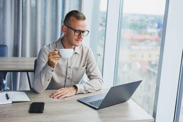 A freelancer in glasses works on a laptop and drinks coffee, a manager sits at a table in the office, works on a laptop. Freelancer workplace, employee at remote work.