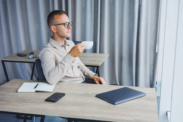 A freelancer in glasses works on a laptop and drinks coffee, a manager sits at a table in the office, works on a laptop. Freelancer workplace, employee at remote work.