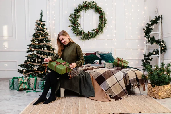 一个年轻的女人在新年的气氛和欢乐中打开圣诞礼物 这个女人在一间明亮舒适的卧室里 布置得很漂亮 以备圣诞节之用 — 图库照片