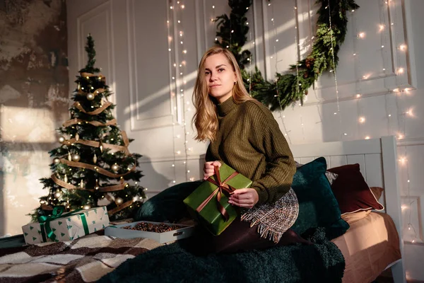 A young woman in a sweater opens Christmas gifts in a bedroom decorated for Christmas against the backdrop of a Christmas tree.