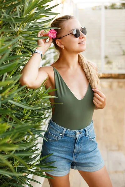 Portrait of a pretty blonde woman in a top and denim shorts, summer vacation, a girl with a pink flower near her face