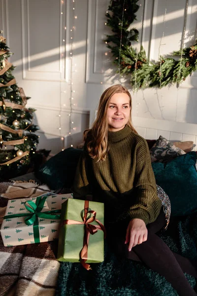 A young woman in a sweater opens Christmas gifts in a bedroom decorated for Christmas against the backdrop of a Christmas tree.