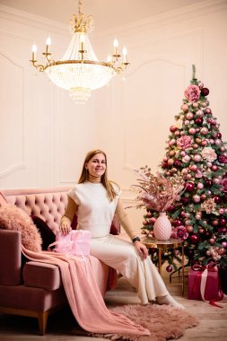Charming woman dressed in a white wool suit holds a pink gift, against the background of a decorated Christmas tree in a room next to a New Year tree and a pink sofa, gifts and candles