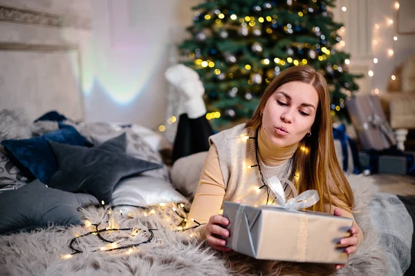 A beautiful blonde woman sits near a Christmas tree in a New Year\'s atmosphere and rejoices. The woman is in a bright, comfortable bedroom, beautifully decorated for Christmas.