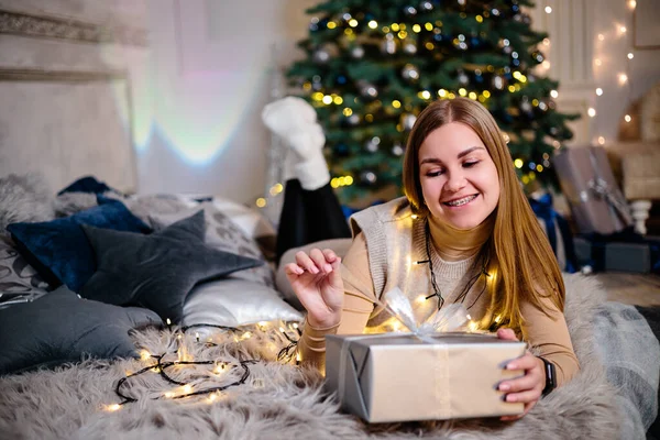 A beautiful blonde woman sits near a Christmas tree in a New Year's atmosphere and rejoices. The woman is in a bright, comfortable bedroom, beautifully decorated for Christmas.