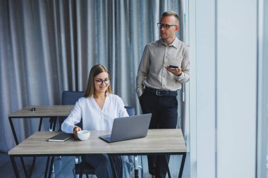 Confident male and female colleagues in elegant casual wear are discussing a project in the workplace, enjoying corporate friendship, happy partners are talking about office networks on a mobile phone