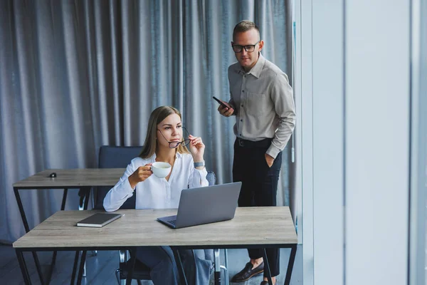 Confident male and female colleagues in elegant casual wear are discussing a project in the workplace, enjoying corporate friendship, happy partners are talking about office networks on a mobile phone