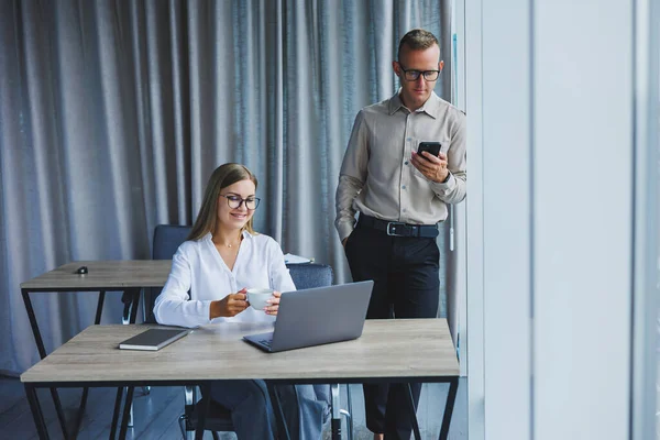 Confident male and female colleagues in elegant casual wear are discussing a project in the workplace, enjoying corporate friendship, happy partners are talking about office networks on a mobile phone