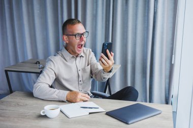 A young attractive male freelancer in glasses sits in the office and looks at the phone. Handsome blogger writing ideas on phone while sitting at wooden table in office with laptop