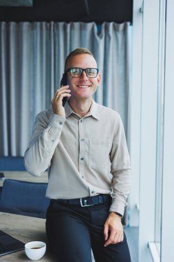 Indoor shot of cheerful bearded male freelancer work at distant, manages finances via smart phone, sits at wooden table, works with documentation, has happy expression. People, work, technology