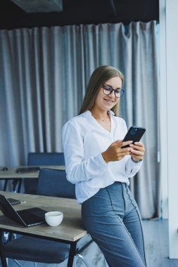 Photo of a successful young woman in a shirt smiling and working on a laptop while talking on the phone in a modern office with large windows. Remote work with coffee