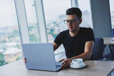 Cheerful businessman in glasses sits at his desk, working with a laptop in the office. A businessman sits and works on a laptop computer and drinks coffee. Starting the day at the office