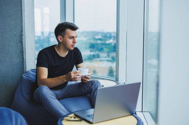Smiling businessman looking for something and typing on a laptop and drinking fresh black coffee. The concept of a modern successful person. Young focused guy with glasses in an open office