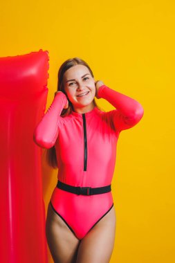 Beautiful young smiling woman wearing a pink swimsuit and posing on a yellow background with an air mattress, isolated on a bright yellow background