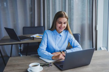A beautiful business woman in a shirt and glasses sits with a laptop in the office at the table. Woman manager in glasses at workplace in modern office. Working day at the computer
