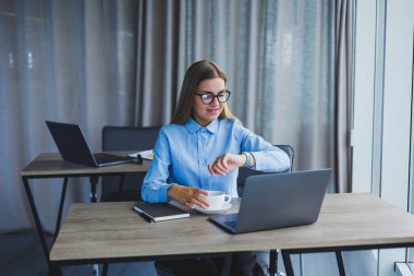 A beautiful business woman in a shirt and glasses sits with a laptop in the office at the table. Woman manager in glasses at workplace in modern office. Working day at the computer