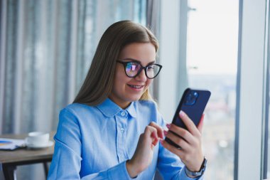 Smart female employee with netbook looking at camera during mobile call, successful freelancer in jacket talking on phone via app