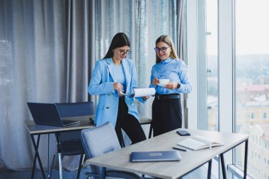 Two young women managers work in a modern office. Women colleagues are talking while working. Friendly staff