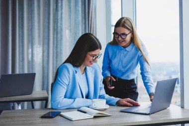 Cheerful employees chatting while working on a laptop, discussing work during a coffee break in the office, a business woman in glasses listens to a colleague, has a pleasant conversation, friendships