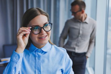 Several friendly colleagues are standing by the office window. A business woman in glasses stands on the background of a man in a shirt. Selective focus. Portrait of business people