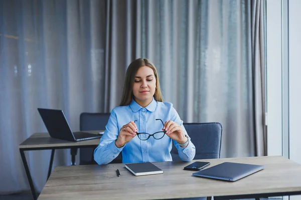 A beautiful woman in glasses sits at a table in the office and works at a laptop. Corporate work in the office. Modern woman workplace manager in classic clothes