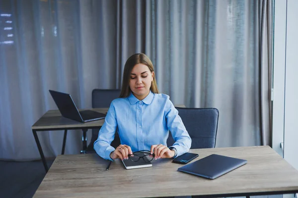 A beautiful woman in glasses sits at a table in the office and works at a laptop. Corporate work in the office. Modern woman workplace manager in classic clothes
