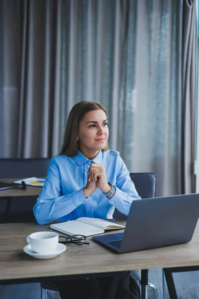 A beautiful business woman in a shirt and glasses sits with a laptop in the office at the table. Woman manager in glasses at workplace in modern office. Working day at the computer