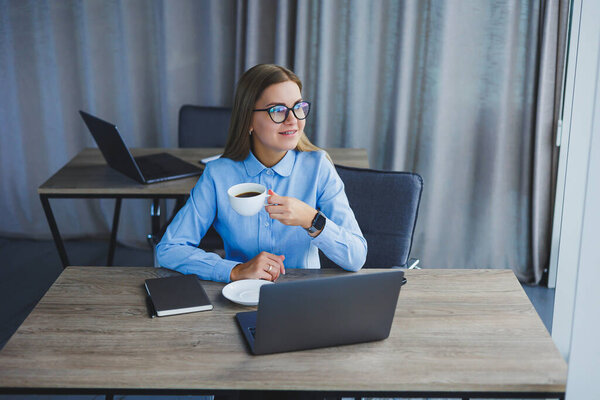 A beautiful business woman in a shirt and glasses sits with a laptop in the office at the table. Woman manager in glasses at workplace in modern office. Working day at the computer