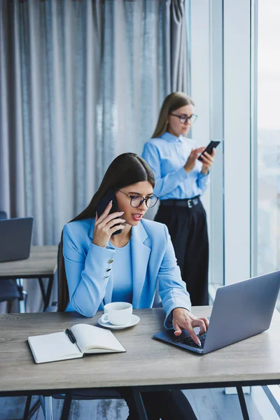 Two women in the office are working and doing business. Business woman in office with glasses and phone. Woman manager in glasses and shirt in the office. Head of working day.