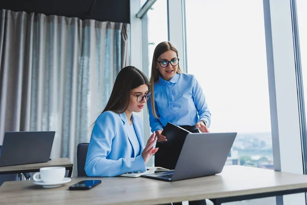 Busy business women with a laptop in the office, watching content on the laptop and discussing the project together, sitting in the office during a working break. Teamwork, corporate discussion