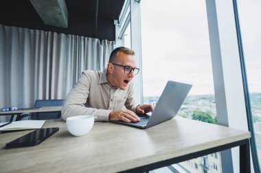 Emotional young manager at work desk with laptop on face glasses. A cheerful businessman is working in a modern office with large windows