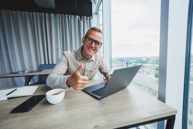 Emotional young manager at work desk with laptop on face glasses. A cheerful businessman is working in a modern office with large windows