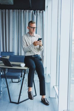 Young smiling businessman holding a cup of coffee in his hands in the office businessman looking out the window standing in a modern workplace man taking a vacation from hard work