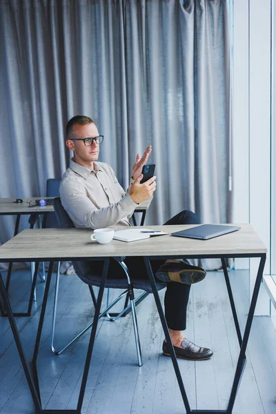 A young attractive male freelancer in glasses sits in the office and looks at the phone. Handsome blogger writing ideas on phone while sitting at wooden table in office with laptop