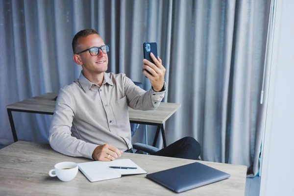 A young attractive male freelancer in glasses sits in the office and looks at the phone. Handsome blogger writing ideas on phone while sitting at wooden table in office with laptop