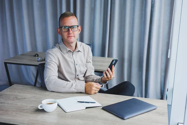 A young attractive male freelancer in glasses sits in the office and looks at the phone. Handsome blogger writing ideas on phone while sitting at wooden table in office with laptop