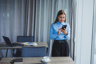 Image of a happy young woman in a jacket smiling and working on a laptop, talking on the phone in a modern office with large windows. Remote work