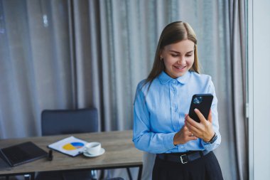 Image of a happy young woman in a jacket smiling and working on a laptop, talking on the phone in a modern office with large windows. Remote work