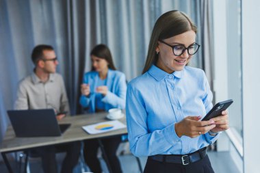Manager woman in classic glasses smiling during working hours in the office talking on the phone, table with laptop, colleagues in the background. A colleague is in the background, selective focus