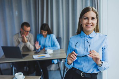 Young business lady. Manager woman in classic glasses smiling during working time in office, desk with laptop, colleagues in background. A colleague is in the background, selective focus