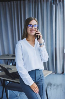 Başarılı, gülümseyen bir iş kadını elinde telefonuyla pencerenin yanında duruyor. Gözlüklü ve beyaz gömlekli bir bayan yönetici telefonda konuşuyor.