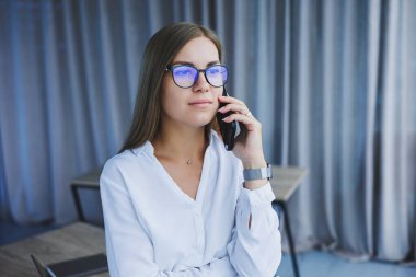 Başarılı, gülümseyen bir iş kadını elinde telefonuyla pencerenin yanında duruyor. Gözlüklü ve beyaz gömlekli bir bayan yönetici telefonda konuşuyor.