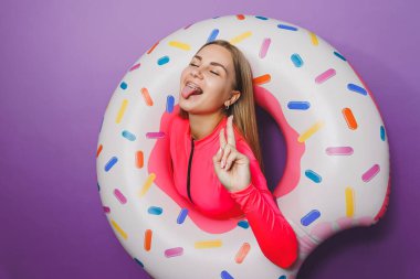 Mor arka planda şişme yüzük tutan uzun saçlı neşeli, ince bir kadın. Pembe mayolu çekici kız.