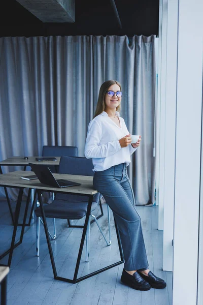 Young smiling business woman standing in office near window with coffee in hands. A female manager in glasses and a white shirt is resting from work